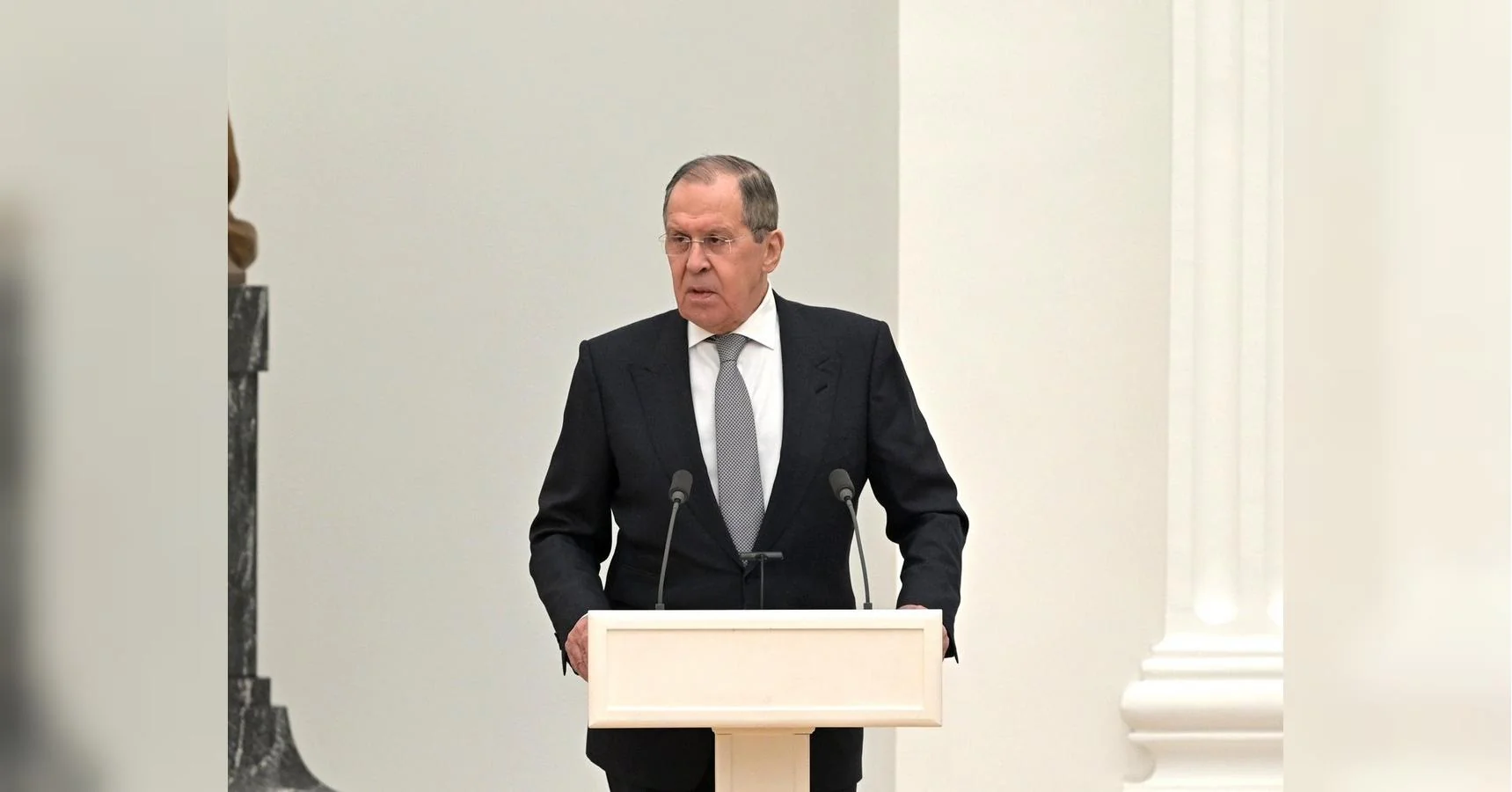 Сергій Лавров у темному костюмі виступає за білою трибуною в залі