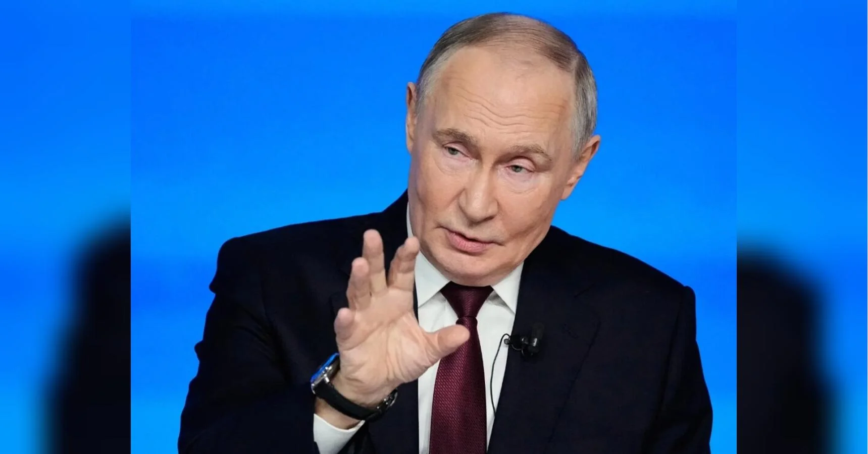 Володимир Путін у костюмі жестикулює рукою під час виступу на синьому тлі