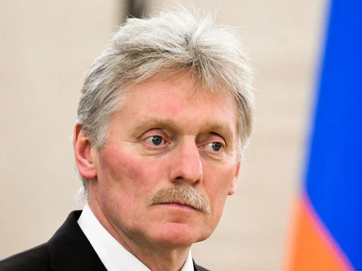 Дмитро Пєсков у костюмі на тлі російського прапора дивиться вбік