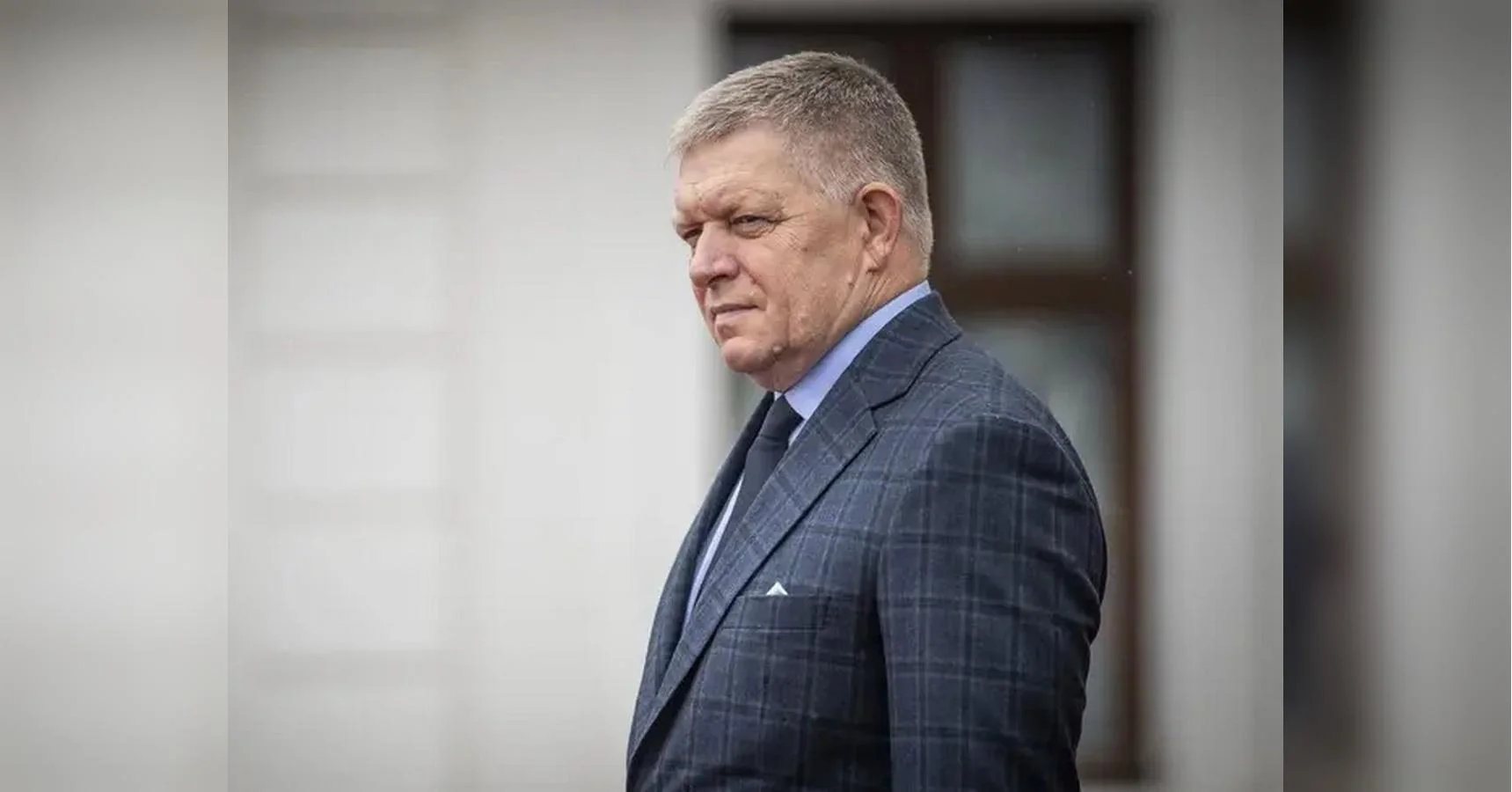 Роберт Фіцо в картатому піджаку стоїть біля вікна світлої будівлі