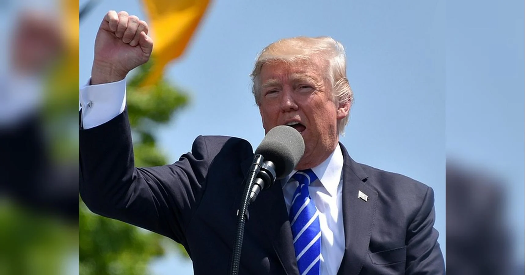 Дональд Трамп у костюмі говорить у мікрофон, піднявши кулак на мітингу