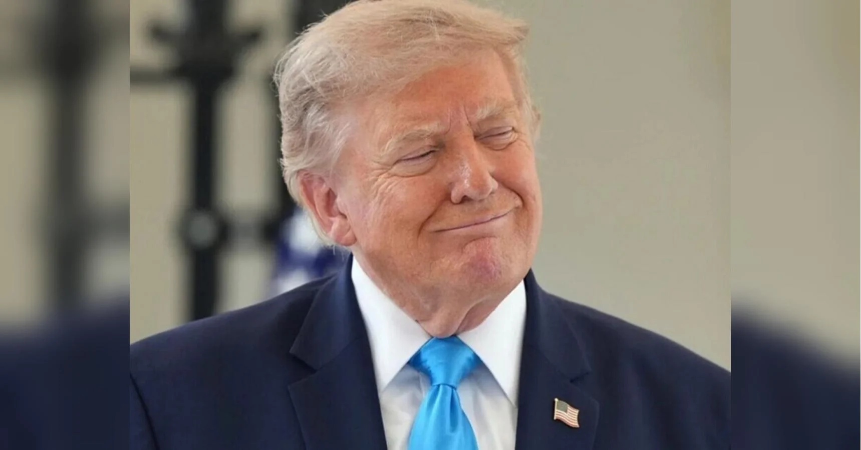 Дональд Трамп у темному костюмі з блакитною краваткою посміхається із заплющеними очима